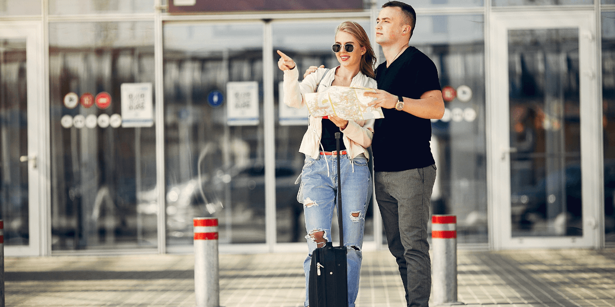 A man and woman stand with luggage in front of a building, ready for travel or check-in.