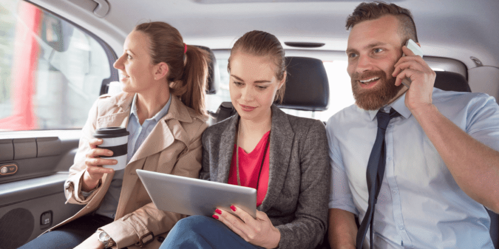 Three professionals riding in a taxi, one holding coffee, one using a tablet, and one talking on the phone.
