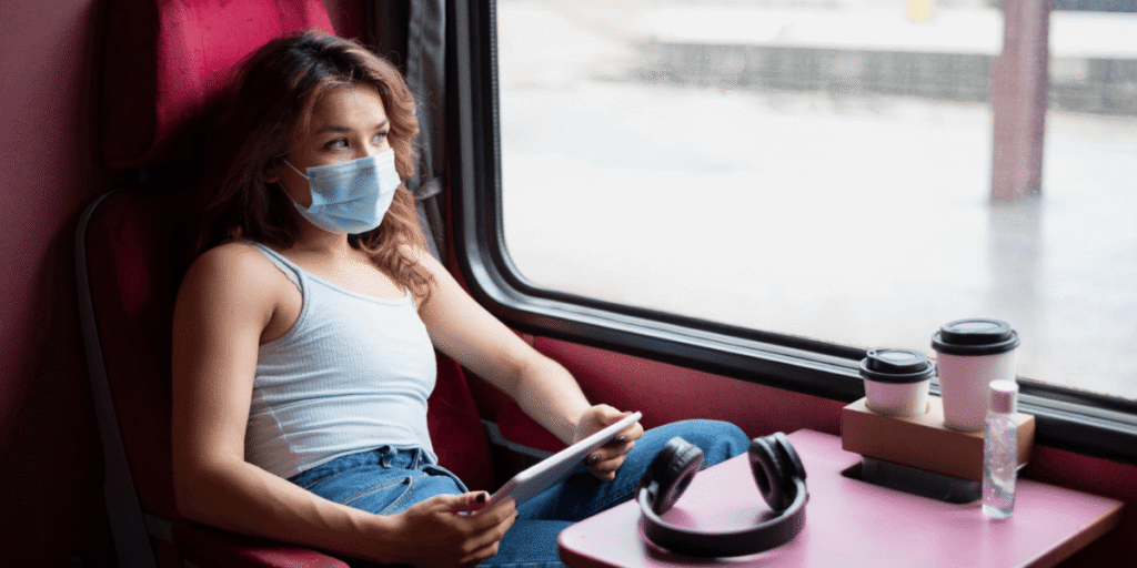 Passenger sitting on a train wearing a mask, holding a tablet with coffee cups and headphones on the table next to her
