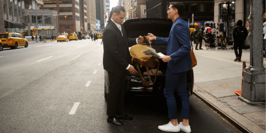 Professional driver assisting a passenger with luggage in front of a luxury car on a city street.