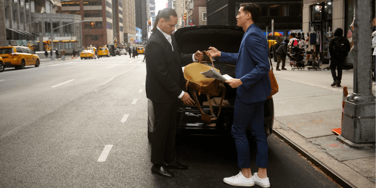 Professional driver assisting a passenger with luggage in front of a luxury car on a city street.