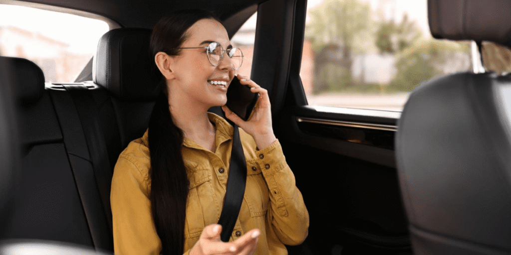Passenger enjoying a phone call while seated in the back of a car, smiling and wearing glasses, with a seatbelt on