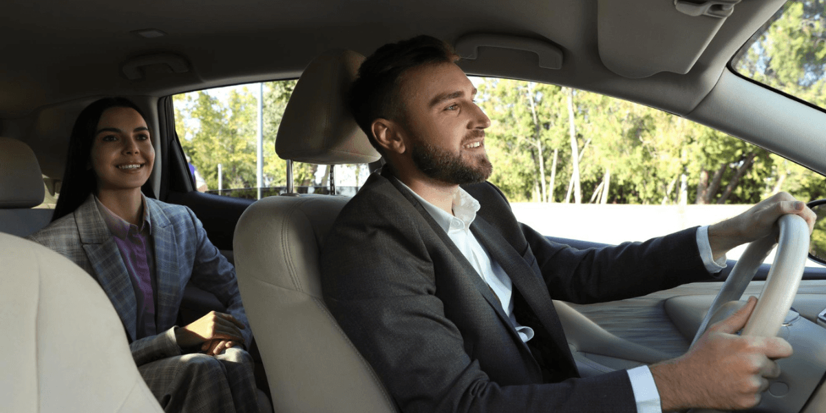 A man driving a car with a woman seated in the back, both dressed in business attire and smiling.