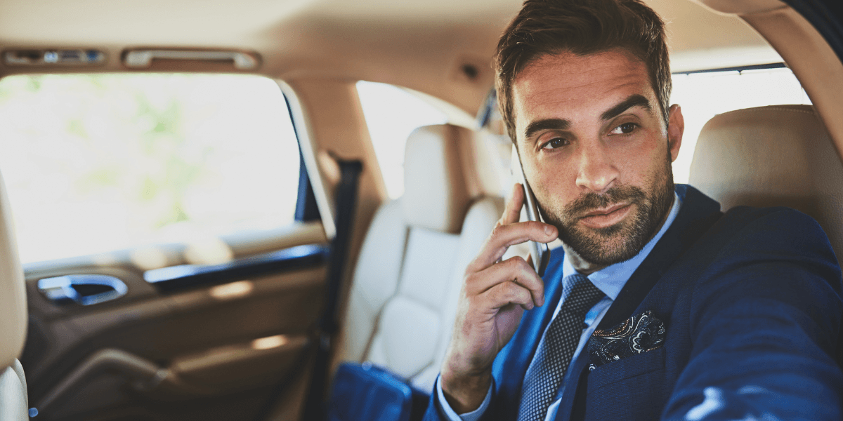 Man in a suit sits in a car's back seat, talking on a phone. He looks out the window with a focused expression, conveying professionalism.