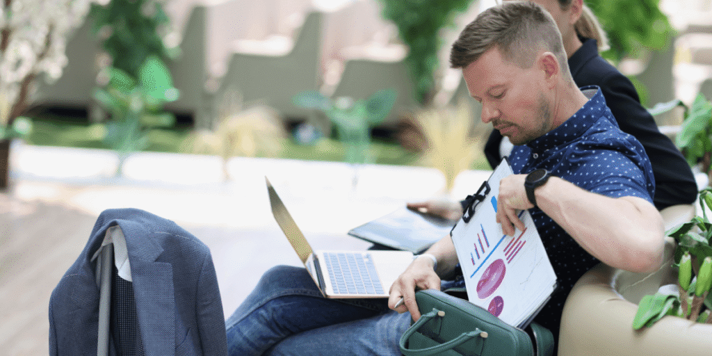 Man sitting casually on a sofa with a laptop, reviewing documents with graphs. He looks focused in a bright, modern setting with plants nearby.