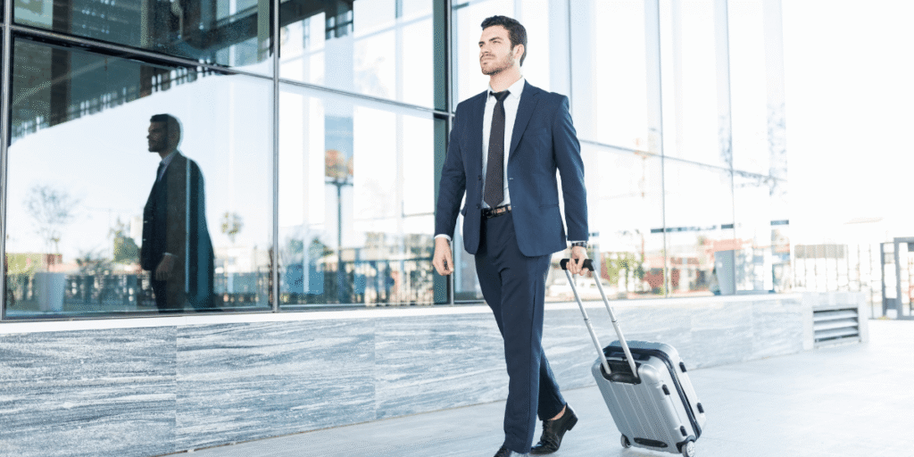 A confident man in a navy suit walks outside a modern glass building, pulling a silver suitcase. The scene conveys a professional and determined tone.