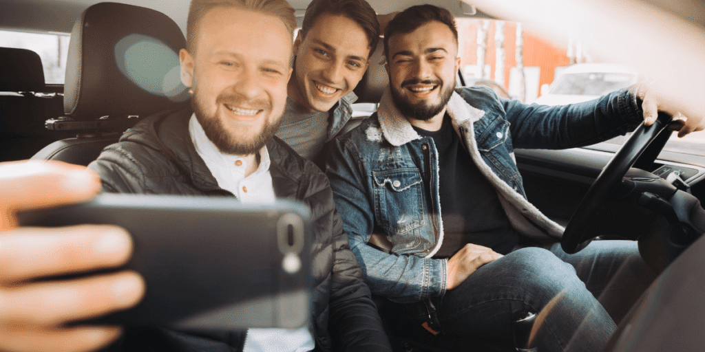 Three friends smiling and taking a selfie inside a car. The mood is cheerful and casual, with natural light illuminating their faces.