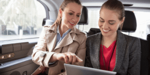 Two women in business attire sit in a car's backseat, smiling and looking at a tablet. One points at the screen, suggesting a collaborative discussion.
