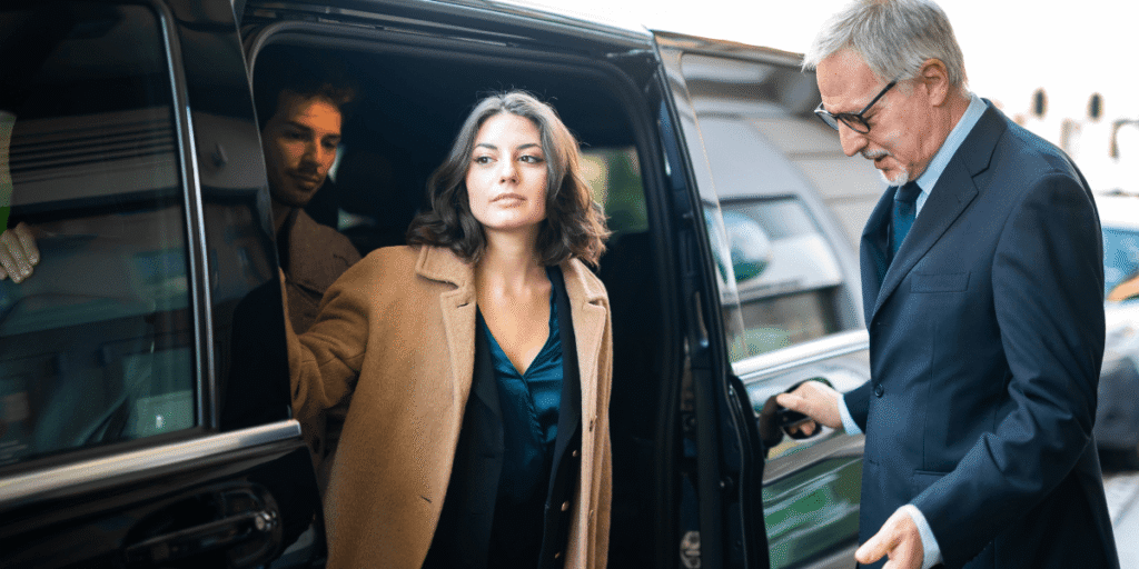 A woman in a tan coat confidently exits a black car, assisted by a driver in a suit. A man is seated inside. The scene conveys professionalism.