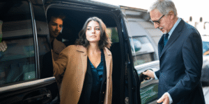A woman in a tan coat confidently exits a black car, assisted by a driver in a suit. A man is seated inside. The scene conveys professionalism.
