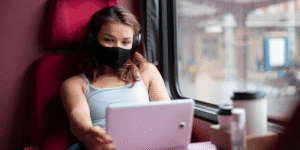 A woman wearing a face mask sits on a train, looking out the window with a thoughtful expression.