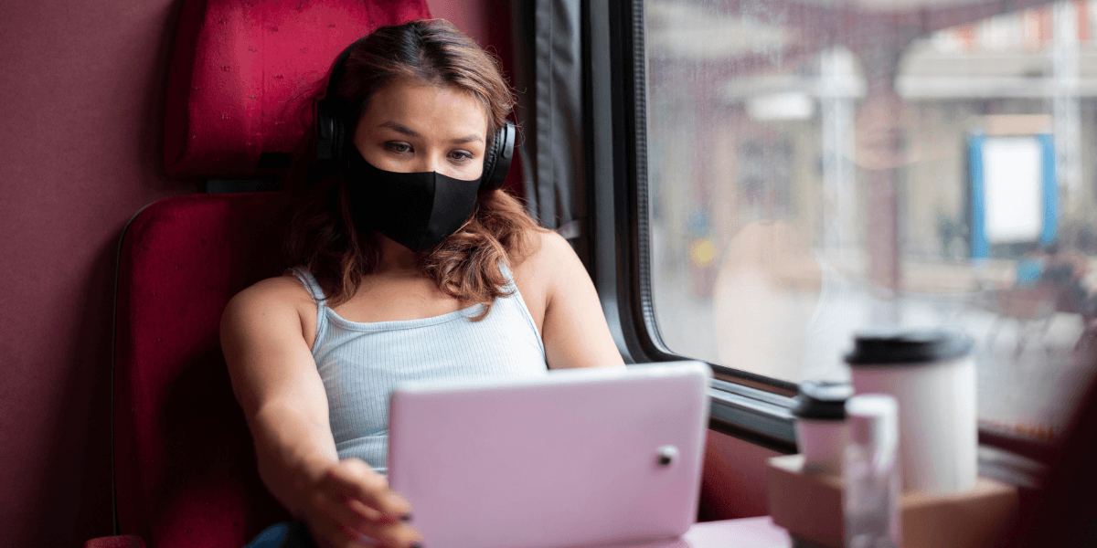 A woman wearing a face mask sits on a train, looking out the window with a thoughtful expression.