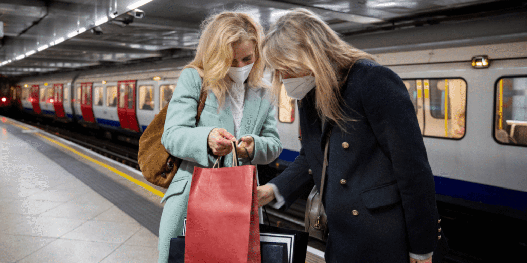Travelling from Leicester to Gatwick Train vs Taxi (1) Two women wearing face masks, smiling while holding colorful shopping bags in a busy outdoor setting.