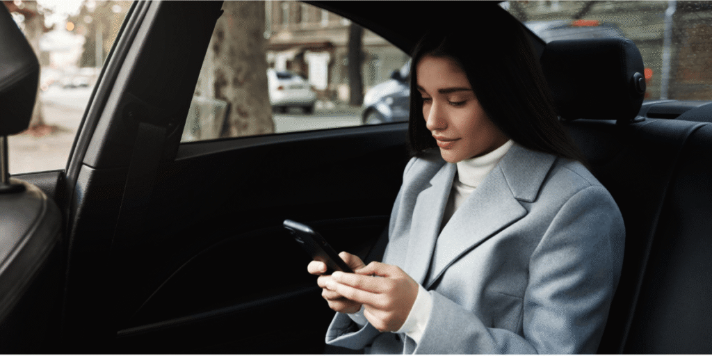 A woman in a light blue coat sits in the back seat of a car, looking down at her smartphone with a neutral expression.