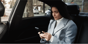 A woman in a light blue coat sits in the back seat of a car, looking down at her smartphone with a neutral expression.