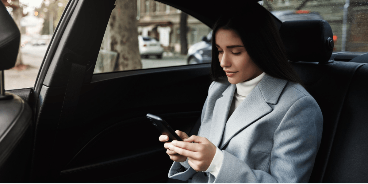 A woman in a light blue coat sits in the back seat of a car, looking down at her smartphone with a neutral expression.