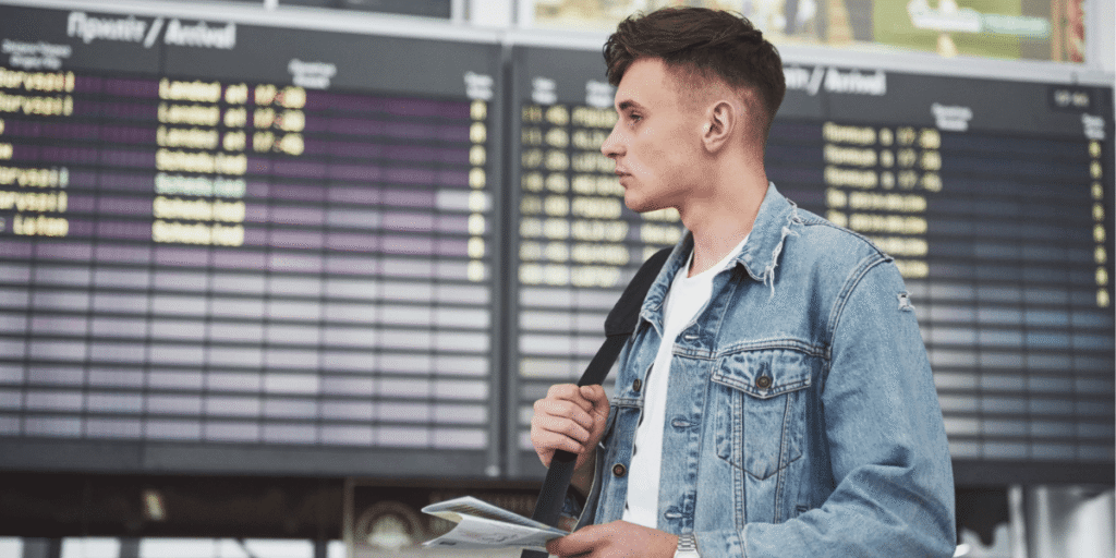 A man wearing a denim jacket stands in front of an airport terminal, ready for travel.