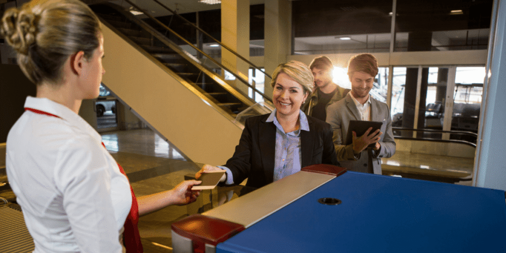 A woman hands a card to a man at an airport, both appearing engaged in conversation.