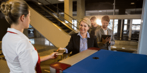 A woman hands a card to a man at an airport, both appearing engaged in conversation.